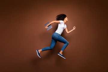 Wall Mural - Full length profile photo of funny dark skin lady jumping high holding notebook hurry enter classroom wear casual white t-shirt jeans isolated brown color background
