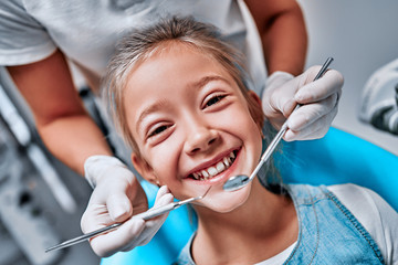 Hands of unrecognizable pediatric dentist making examination procedure for smiling cute little girl sitting on chair in hospital.