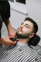 Portrait of a man in barber shop, lying in a chair and looking up while the barber cuts his beard. Barber trims his beard seriously to the client. Vertical closeup portrait.