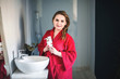 © algae5 - young woman in a pink terry bathrobe is standing in the bathroom smiling and holding jars of makeup in her hands