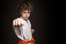 Karate Kid Fist On Dark Background. Caucasian Boy Keeping A Karate Position Wearing The Kimono And The Orange Belt