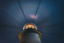 The Sea Lighthouse Shines At Night With Bright Rays Of Light Against The Background Of The Starry Sky