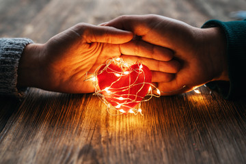 Wall Mural - Two hands holding a red heart shape covered with led lights on wooden and bokeh lights background. Valentines day and romance concept.