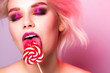 © Yuliya Timofeeva - A model with bright makeup licks a candy on a pink background with eyes closed.