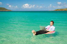 Barefoot businessman working on his laptop while floating on a colorful inflatable ring in bright tropical waters