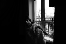A Young Woman Wrapped In A Blanket Sitting On A Chair With A Glass Of Red Wine Near The Big Window With A Metal Fence. Black And White Photo