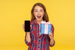 © khosrork - Amazed shocked excited ginger girl in shirt standing with wrapped gift box and mobile phone in her hands, holding present, looking with surprised face. indoor studio shot isolated on yellow background