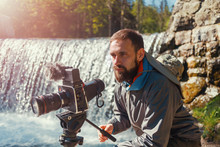 Travel Photographer Bearded Man Close-up With Professional Film Camera On Tripod Shooting Mountain Landscape In Waterfall Background. Hiker Tourist Professional Photography Shooting, Filming Backstage