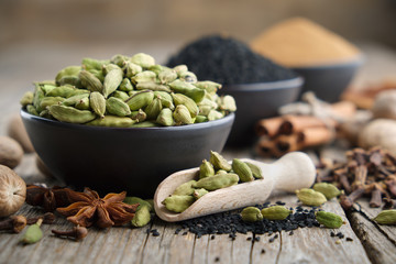 Green cardamom pods in black ceramic bowl in the foreground. Aromatic spices: anise, gloves, black cumin seeds, nutmegs, cinnamon sticks, turmeric. Ingredients for cooking. Ayurveda treatments.