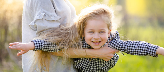 A young mother shakes her little daughter in her arms in the spring park. The girl spread her arms and flies like a plane in her mother’s arms.