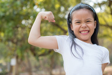 Happy Little Asian Girl Child Standing Showing Front Teeth With Big Smile.  Showing Arms Muscles Smiling Proud. Looking Camera Showing Biceps. Fresh Healthy Green Bio Background. Fitness Concept.