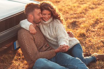 Wall Mural - Beautiful young couple enjoying time together outdoor sitting on meadow leaning on old fashioned car embracing each other.