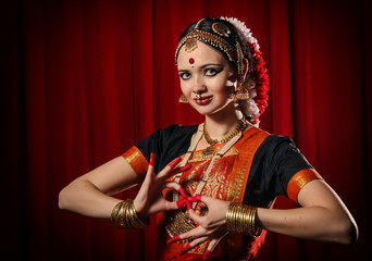 Portrait of white girl as an Indian classical dancer in traditional dress and performing dance performance on the red curtain background. Classical indian temple dance form Bharatanatyam. Dance pose