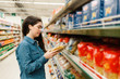 © _KUBE_ - Shopping at the grocery store. A young woman in a denim shirt reads the information on a packet of pasta. In the foreground blurred shelves with products. In profile