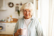 © shurkin_son - Positive friendy looking senior elderly woman with gray hair and wrinkles spending day at home, posing by window with cozy clean kitchen in background, drinking tea or coffee in the morning