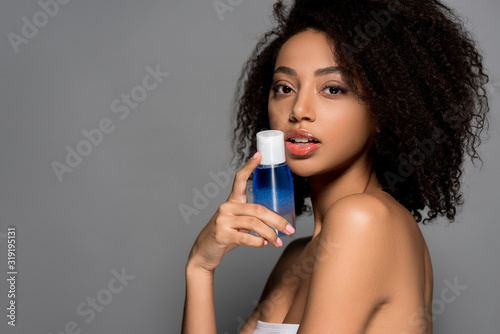 sexy african american girl holding bottle of makeup remover, isolated on grey