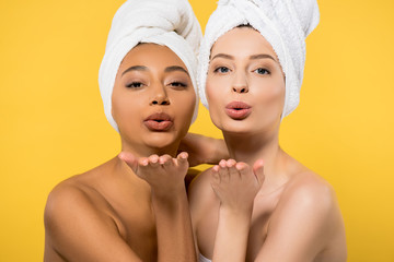 Wall Mural - positive multiethnic girls with towels on heads blowing air kisses, isolated on yellow