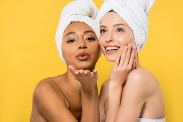 Wall Mural - happy multicultural young woman with towels on heads blowing air kiss, isolated on yellow