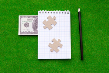 Notepad, two puzzle pieces, American dollar bill, pencil on a green background. Business planning, Finance.