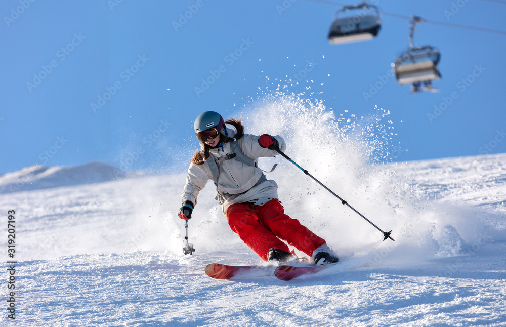 Girl On the Ski. a skier in a bright suit and outfit with long pigtails on her head rides on the track with swirls of fresh snow. Active winter holidays, skiing downhill in sunny day. Woman skier - obrazy, fototapety, plakaty 