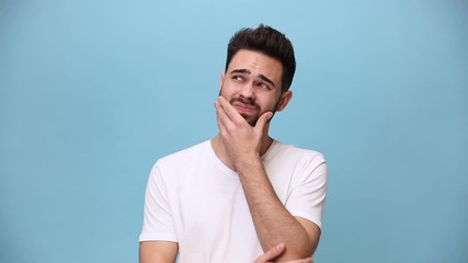 Wall Mural - Handsome unshaven bearded young guy 20s wearing white t-shirt isolated on pastel blue background in studio. People sincere emotions lifestyle concept Look at camera pensive thoughtful put hand on face