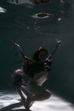 Beautiful Girl Swims Underwater In The Pool With A Scarf Or In A Dress. Brunette With Dark And Long Hair. Dark Tones Of Water And Atmosphere
