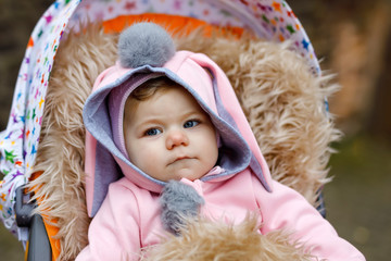 Cute little beautiful baby girl sitting in the pram or stroller on autumn day. Happy smiling child in warm clothes, fashion stylish pink baby coat with bunny ears. Baby going on a walk with parents.