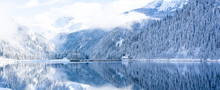 Panoramic View Of Beautiful White Winter Wonderland Forest Scenery In The Alps With Snowy Mountain Summits Reflecting In Crystal Clear Mountain Lake On A Cold Sunny Day With Blue Sky And Clouds