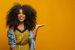 © Brastock Images - Cheerful Afro woman points away on copy space, discusses amazing promo, gives way or direction, wears yellow warm sweater, has pleasant smile, feels optimistic, isolated over yellow background.