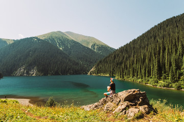 Kazakhstan, Kolsai Lake. The second Kolsai Lake in the mountains among pine forests in the sunlight. Turquoise water and yellow grass A blonde girl in shorts is standing by the lake