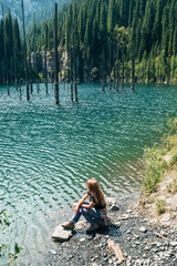 Wall Mural - Kazakhstan, Lake Kaindy. Lake with turquoise water. There are dead standing trunks of old trees in the lake. Dead forest in the water. A girl with her hair sits on the shore of Lake Kaindy.