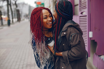 two beautiful and stylish dark-skinned girls with long hair standing in a autumn city near purple wall and drinking a coffee