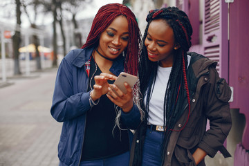 two beautiful and stylish dark-skinned girls with long hair standing in a autumn city near purple wall and using the phone