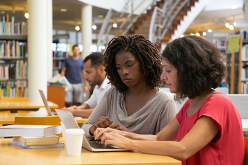 Two thoughtful women talking and typing on laptop. Concentrated ladies talking while sitting at table with modern device at public library. Education, technology concept