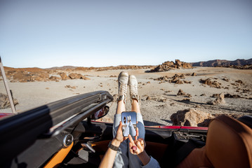 Wall Mural - Young woman traveling by convertible car on the desert valley, pulling legs out the window and photographing with phone