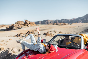 Wall Mural - Lifestyle portrait of a young woman enjoying road trip on the desert valley, lying on the car hood and photographing on phone