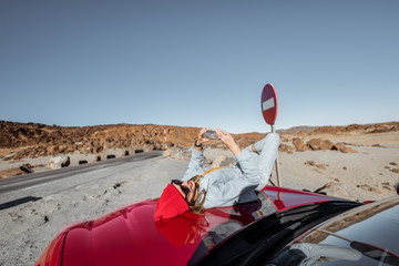 Wall Mural - Lifestyle portrait of a young woman enjoying road trip on the desert valley, lying on the car hood and photographing on phone