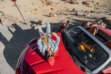 Wall Mural - Lifestyle portrait of a young woman enjoying road trip on the desert valley, lying on the car hood and photographing on phone. View from above