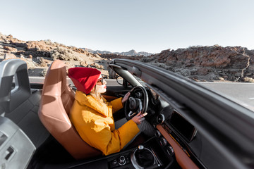 Wall Mural - Happy woman in bright hat and jacket driving convertible car while traveling on the desert road on a sunset. Image focused on the road, woman is out of focus