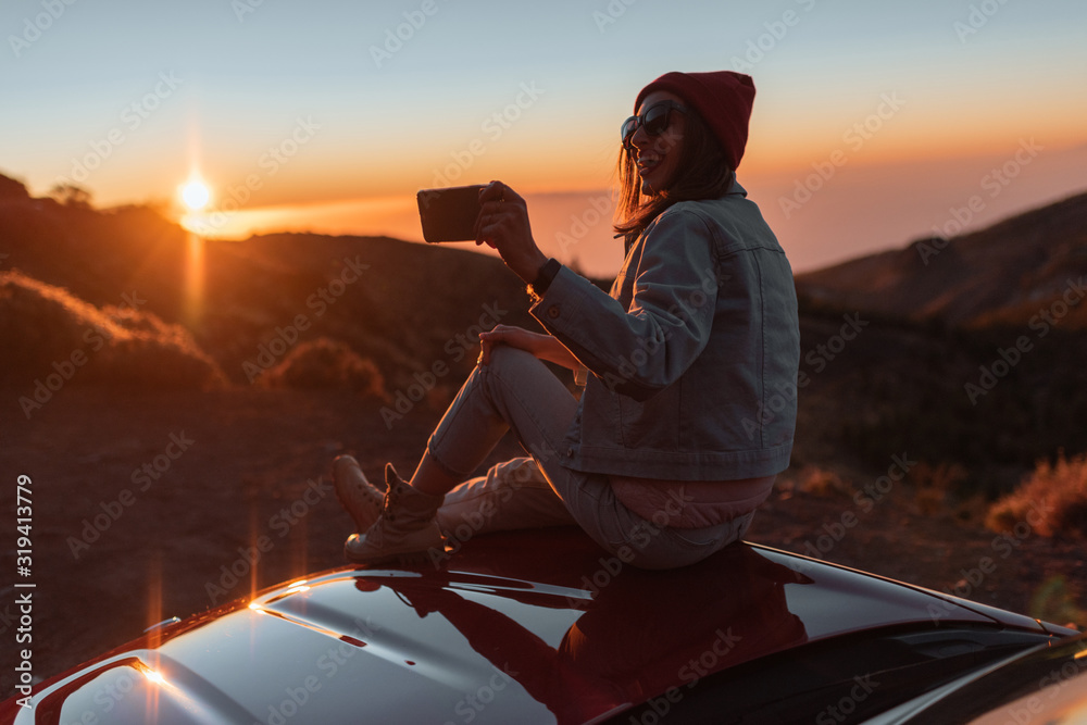 Young woman photographing with phone beautiful landscape during a sunset, sitting on the car hood while traveling high in the mountains - obrazy, fototapety, plakaty 