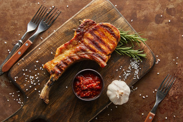 Wall Mural - Top view of ribeye steak with tomato sauce and garlic on chopping board by forks on stone background