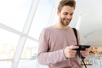 Canvas Print - Image of young bearded man holding smartphone over bright window indoors