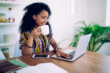 Cheerful young modern woman browsing laptop while drinking coffee at home