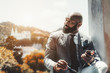 © skyNext - A handsome mature bearded African man in a formal suit with a cellphone and eyeglasses in his hands; an adult elegant bald black guy with a beard is using his smartphone outdoors, landscape behind him