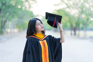 Portrait of a young cheerful female graduate wearing academic gown holding graduate cap celebrate her university degree in commence day in college campus.