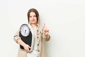 Wall Mural - Young curvy woman holding a scale showing number one with finger.