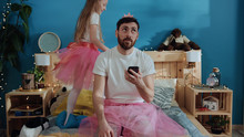 Portrait Of Tired Bored Young Man In Fairy Dress Using A Smartphone Sitting On Bed. Happy Princess Girl Dancing Around Waving Magic Wand Playing With Her Daddy At Home.