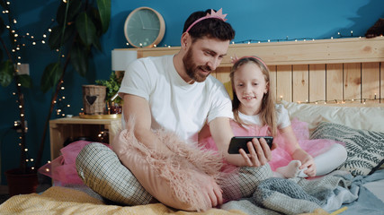 Wall Mural - Family portrait of young daddy and his daughter dressed like fairies watching funny cartoons on smartphone sitting together in bedroom. Exciting family party. Joy and fun.