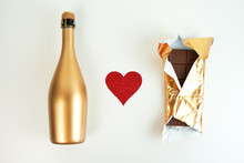 Golden Champagne Bottle And Chocolate Bar In Gold Wrapper And Red Glitter Heart On A White Background. Festive Set. Top View