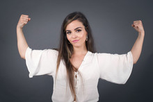 Waist Up Shot Of Caucasian Woman Raises Arms To Show Her Muscles Feels Confident In Victory, Looks Strong And Independent, Smiles Positively At Camera, Stands Against Gray Background. Sport Concept.